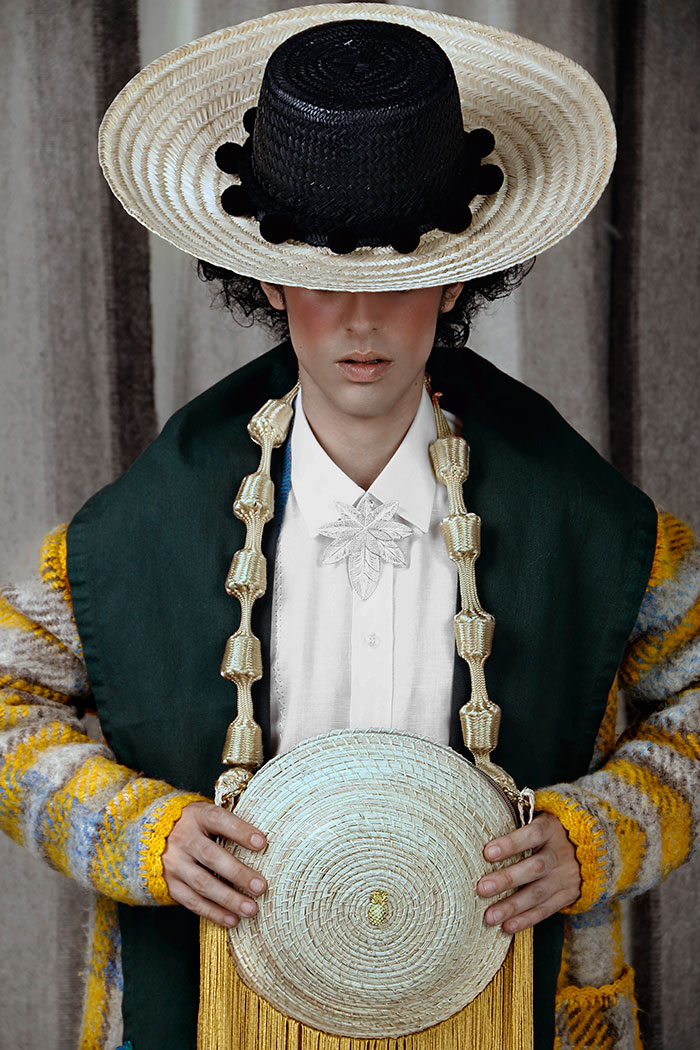 Tomás lleva un sombrero Wayúu elaborado por el artesano Gary González; con un gabán de lana y seda de Edue Marcial Pérez sobre una guayabera de lino bordada a mano en Cartago, de Eliseo Gutiérrez; complementada con broche de filigrana del maestro Eligio Rojas y poncho de Adriana Santacruz, de Pasto. El bolso, elaborado en palma de iraca y flecos de acrílico con pompones forrados en hilo de seda, es de Palma Canaria. Jean negro de Orozco Clothing; zapatos charolados de Dr. Martens. Chivos tejidos en cabuya por artesanos de Santander en el Laboratorio de Innovación y Diseño de Artesanías de Colombia.